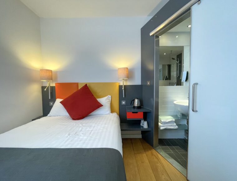 Compact double room with a double bed, bedside table and bathroom separated with a sliding door.